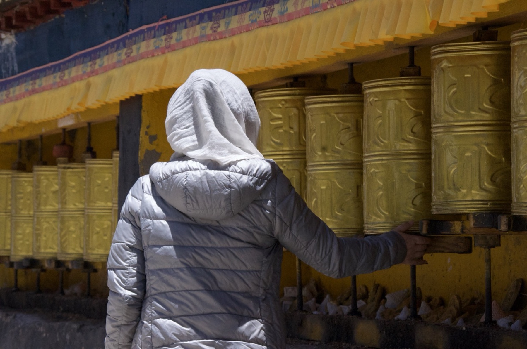 A hooded figure rests a hand on the base of a prayer wheel in a long golden row.